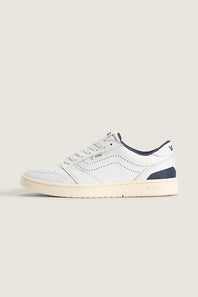 A low-top sneaker in white leather with navy blue accents on the collar, heel, and tongue, featuring a cream-colored rubber sole, shown against a solid light beige background.
