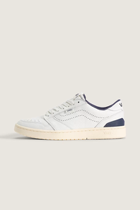 A low-top sneaker in white leather with navy blue accents on the collar, heel, and tongue, featuring a cream-colored rubber sole, shown against a solid light beige background.