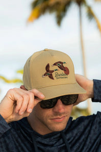 A man wearing a tan snapback cap with an embroidered duck logo and the text 'n the front, photographed outdoors with a blurred tropical background.