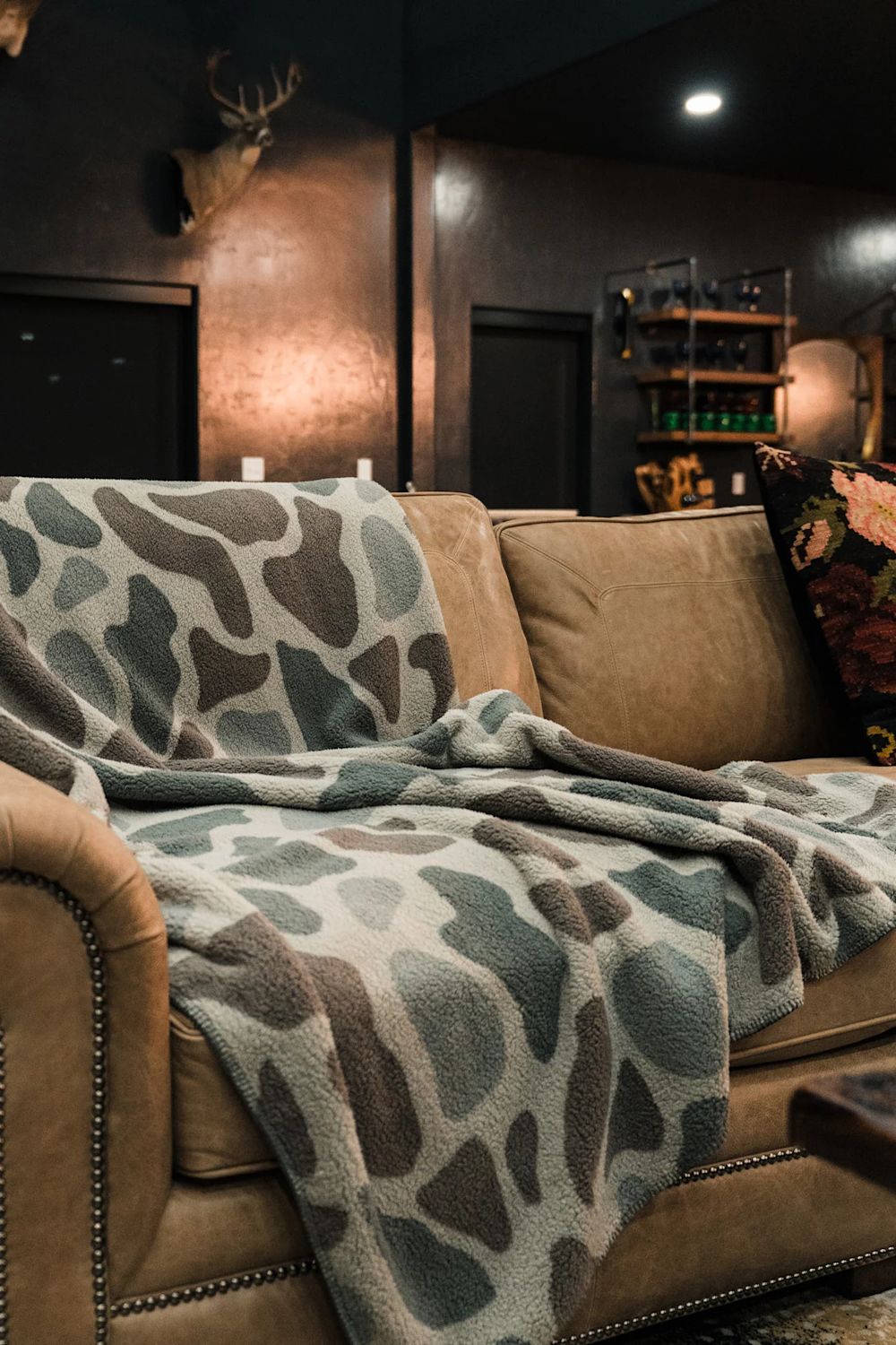 A cozy brown leather sofa in a dimly lit room, draped with a plush throw blanket featuring a gray, beige, and brown organic spot pattern.