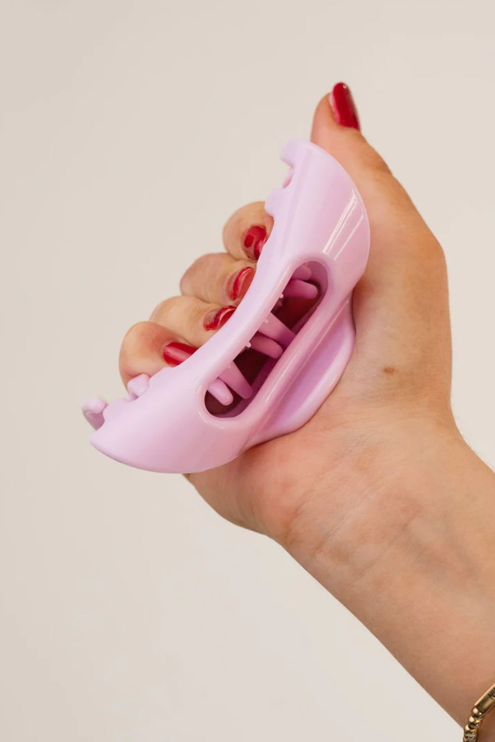 A hand with red painted fingernails holds a light pink silicone nail file or cuticle pusher against a plain off-white background.