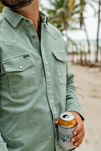 A man wearing a sage green western-style button-down shirt holds a can of beer in a white can cooler with a black and green 'Poncho' logo, set against a blurred beach background with palm trees.