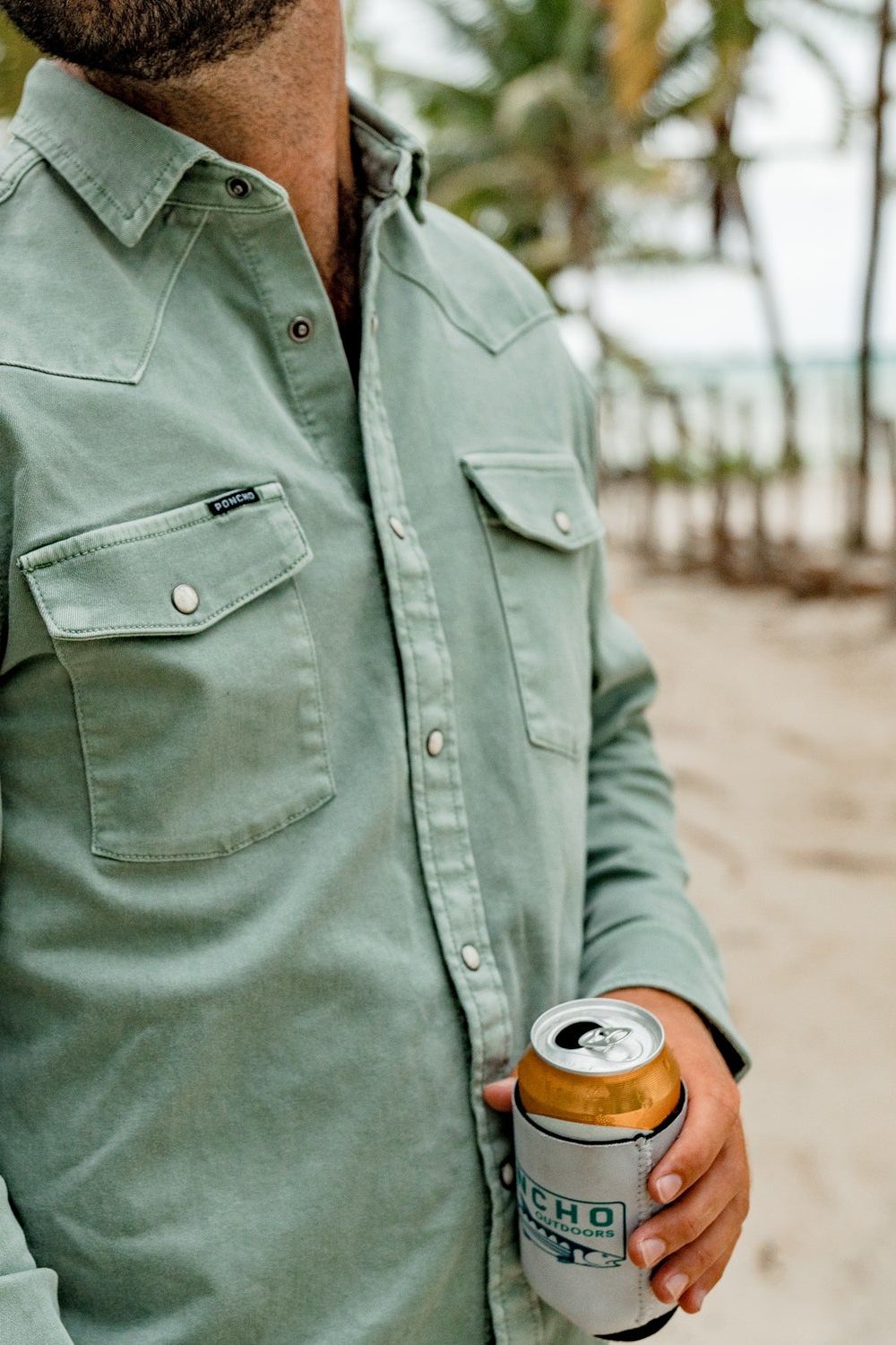 A man wearing a sage green western-style button-down shirt holds a can of beer in a white can cooler with a black and green 'Poncho' logo, set against a blurred beach background with palm trees.