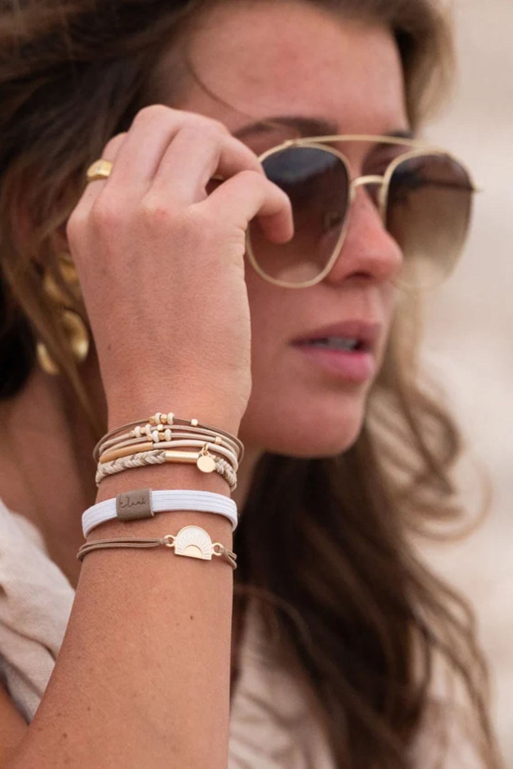 A close-up lifestyle shot of a woman wearing gold-framed aviator sunglasses and multiple delicate bracelets on her wrist, including a white band with a gold heart charm and a small gold tag.