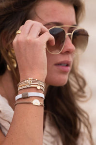 A close-up lifestyle shot of a woman wearing gold-framed aviator sunglasses and multiple delicate bracelets on her wrist, including a white band with a gold heart charm and a small gold tag.