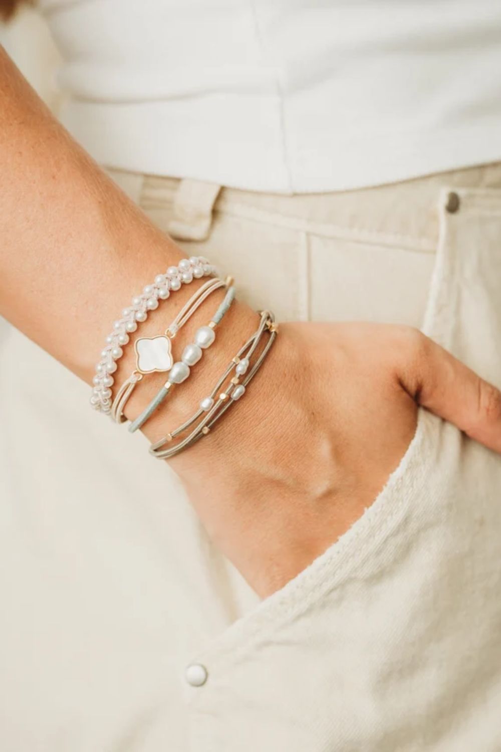 A close-up of a person's wrist wearing a stack of delicate bracelets, including pearl strands, a white shell bracelet, and thin silver and rose gold bangles, paired with a white top and beige pants.