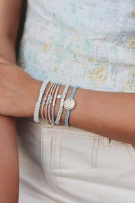 A close-up of a person's wrist wearing multiple delicate bracelets, including silver-toned wire cuffs, a white textured band, a light blue cord bracelet, and a round silver charm bracelet, paired with a light patterned top and cream pants.