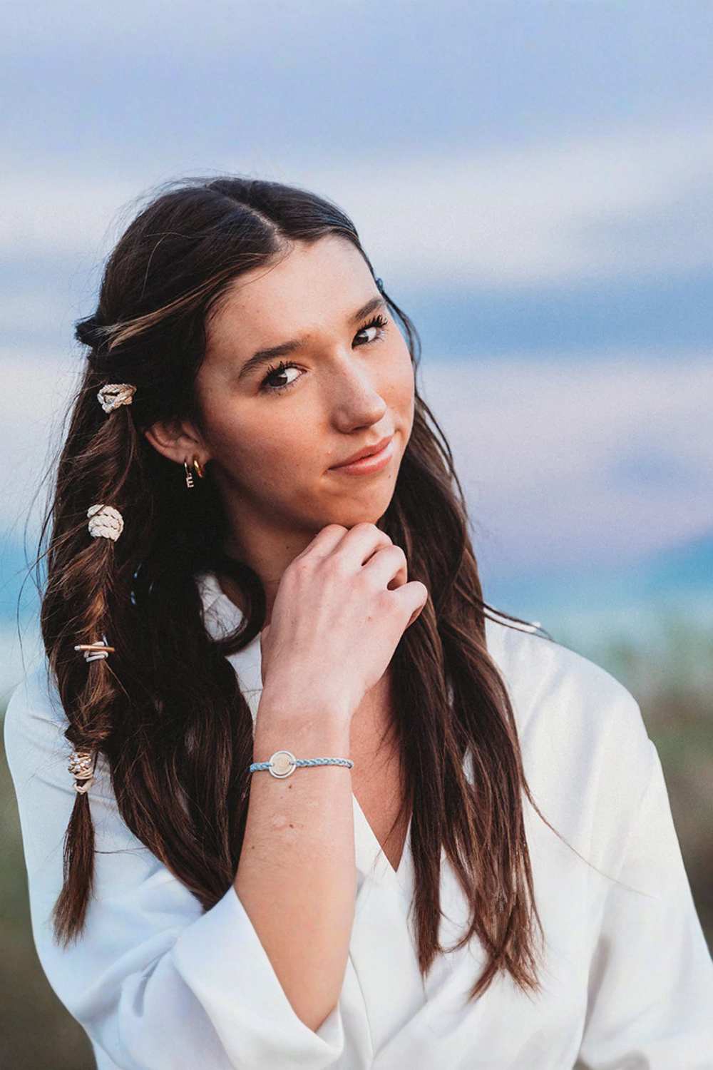 A woman with long dark brown hair styled in a half-up braid with decorative hair clips, wearing a white blouse and a delicate silver bracelet, photographed outdoors against a soft-focus sky background.