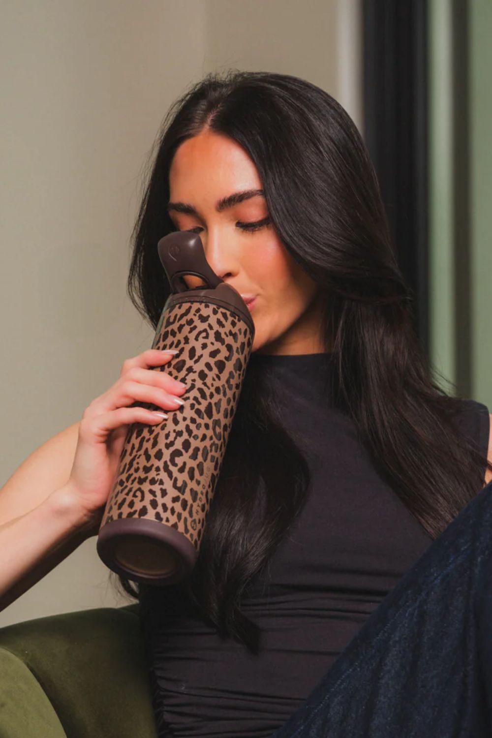 A woman with long dark hair holds a leopard-print insulated tumbler to her lips while wearing a black top in a softly lit indoor setting.