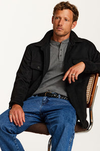 A man wearing a black overshirt, gray henley shirt, and blue jeans sits on a chair against a plain white background.