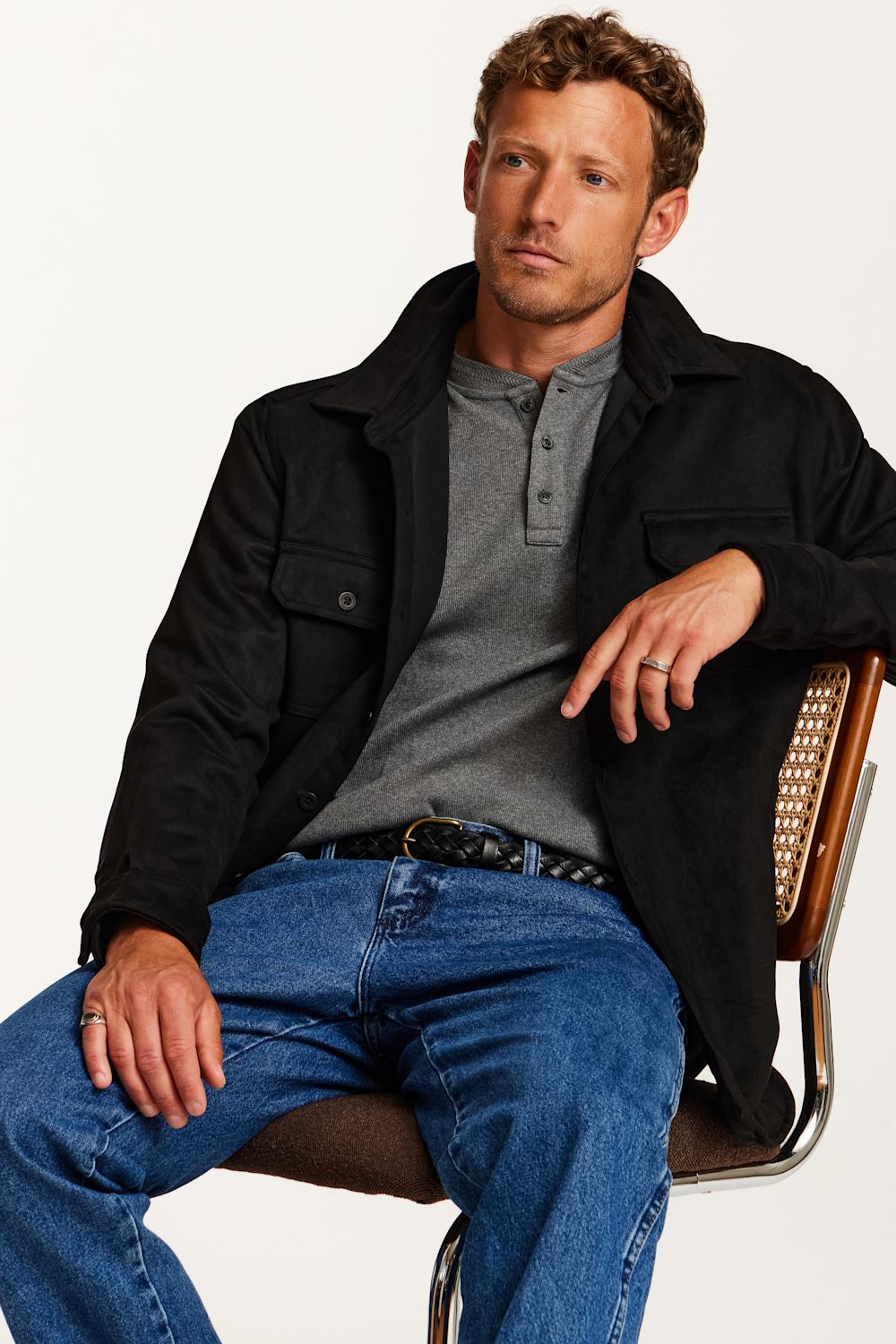 A man wearing a black overshirt, gray henley shirt, and blue jeans sits on a chair against a plain white background.