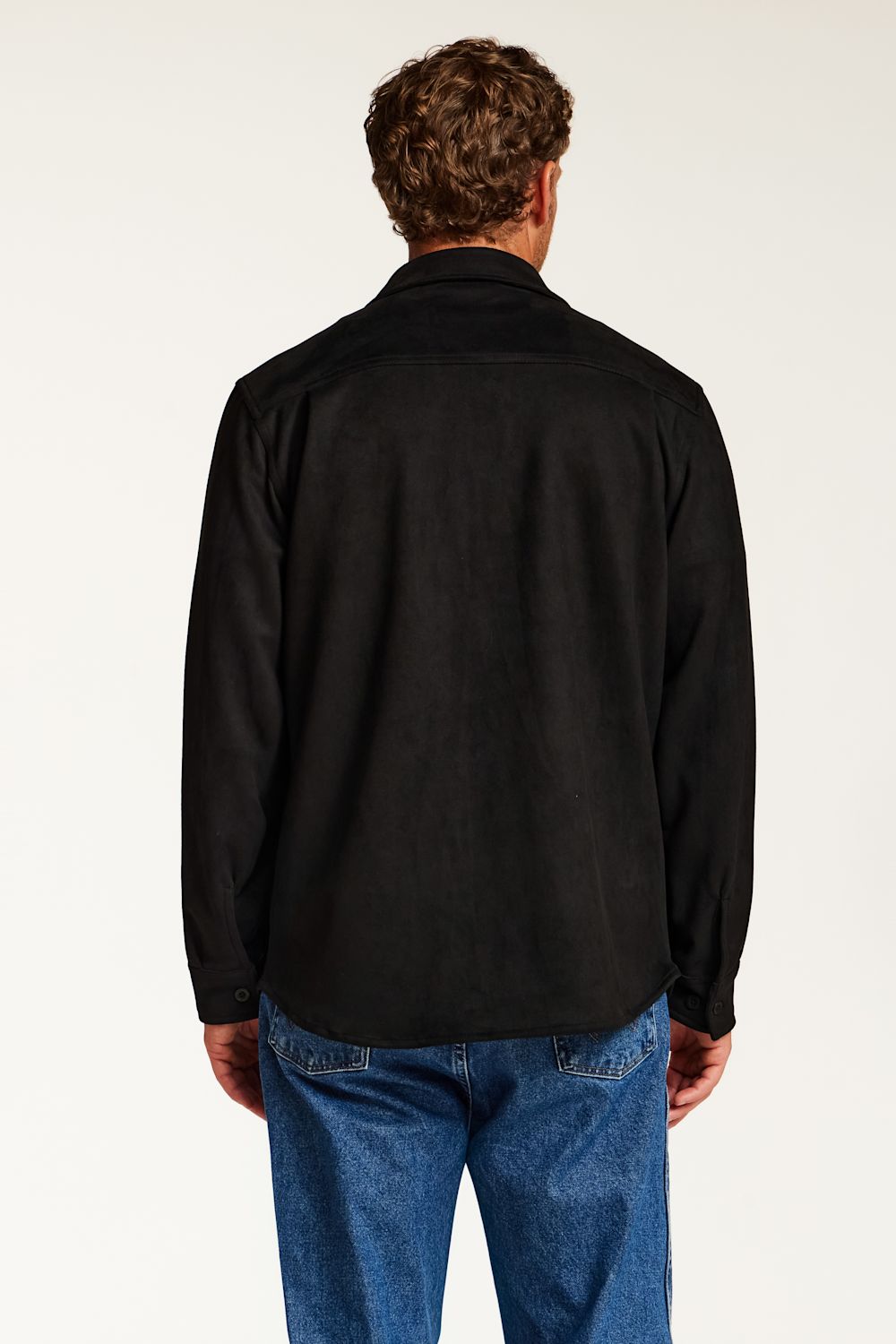 A man viewed from behind wearing a black long-sleeve button-up shirt and blue jeans against a plain white studio background.