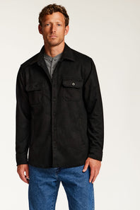 A man wearing a black button-up overshirt with two chest pockets over a gray t-shirt and blue jeans, standing against a plain light background.