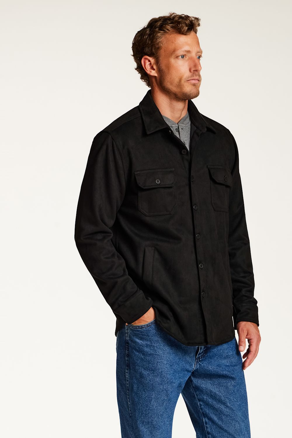 A man wearing a black long-sleeve button-up shirt jacket over a gray shirt and blue jeans, standing against a plain white studio background.