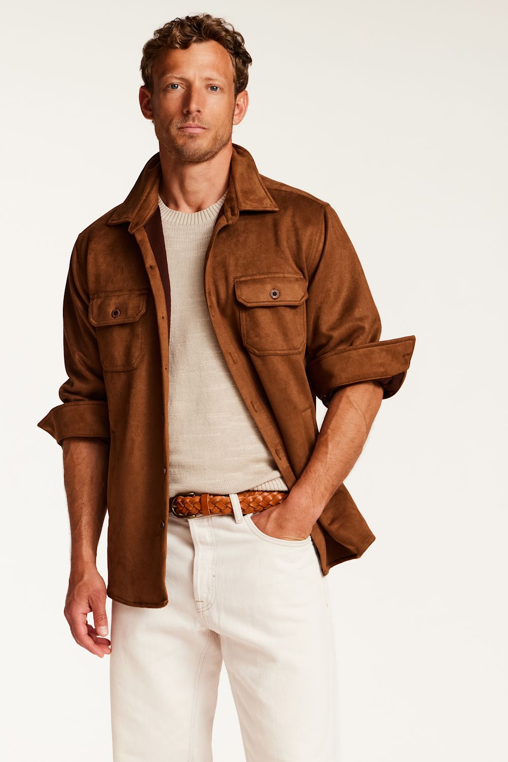 A man wearing a brown suede-like button-up shirt jacket over a cream knit sweater, white trousers, and a brown woven belt, photographed against a plain light background.