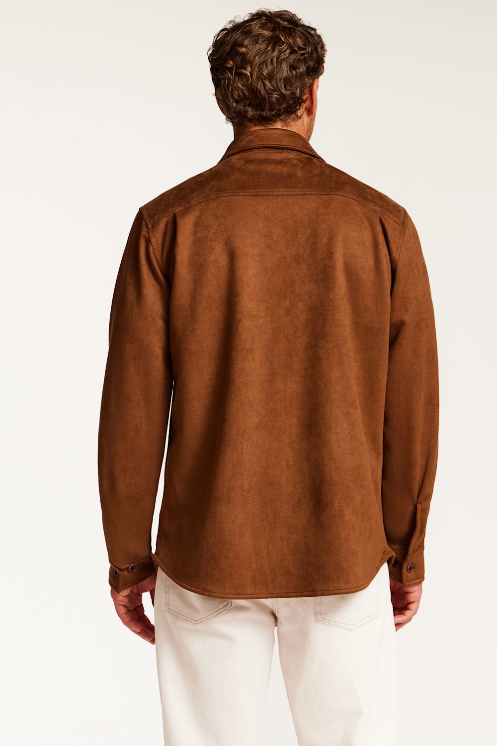 A man viewed from behind wearing a brown suede or faux-suede long-sleeve shirt and off-white trousers, against a plain white studio background.