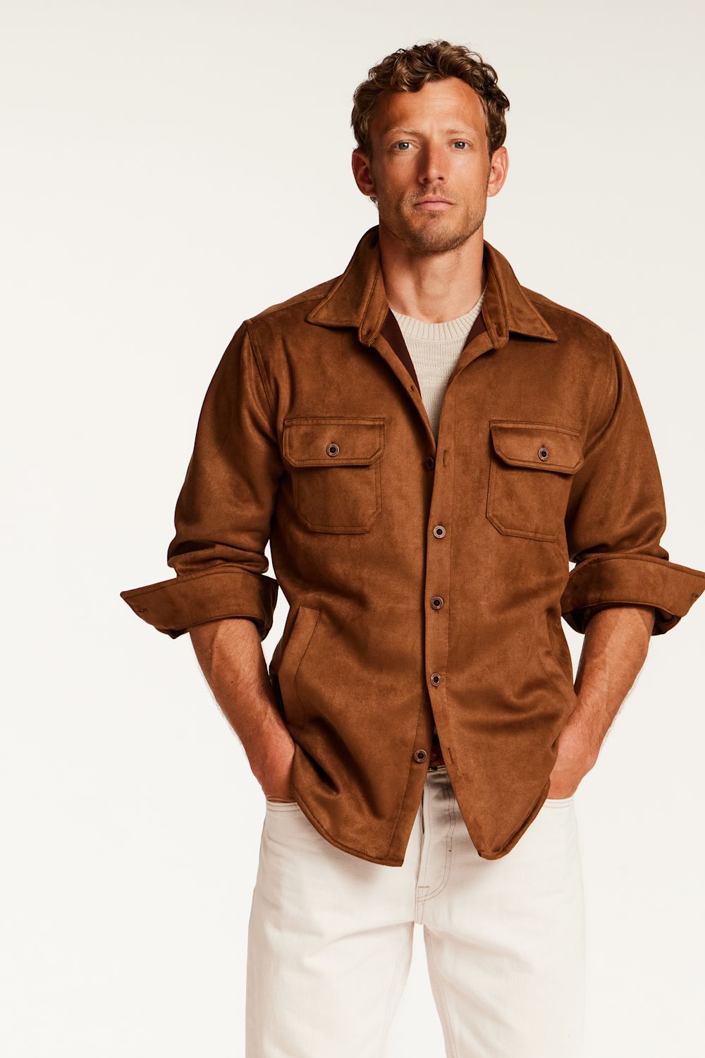 A man wearing a brown suede-like button-up shirt jacket over a beige knit sweater and white pants, standing against a plain light background.
