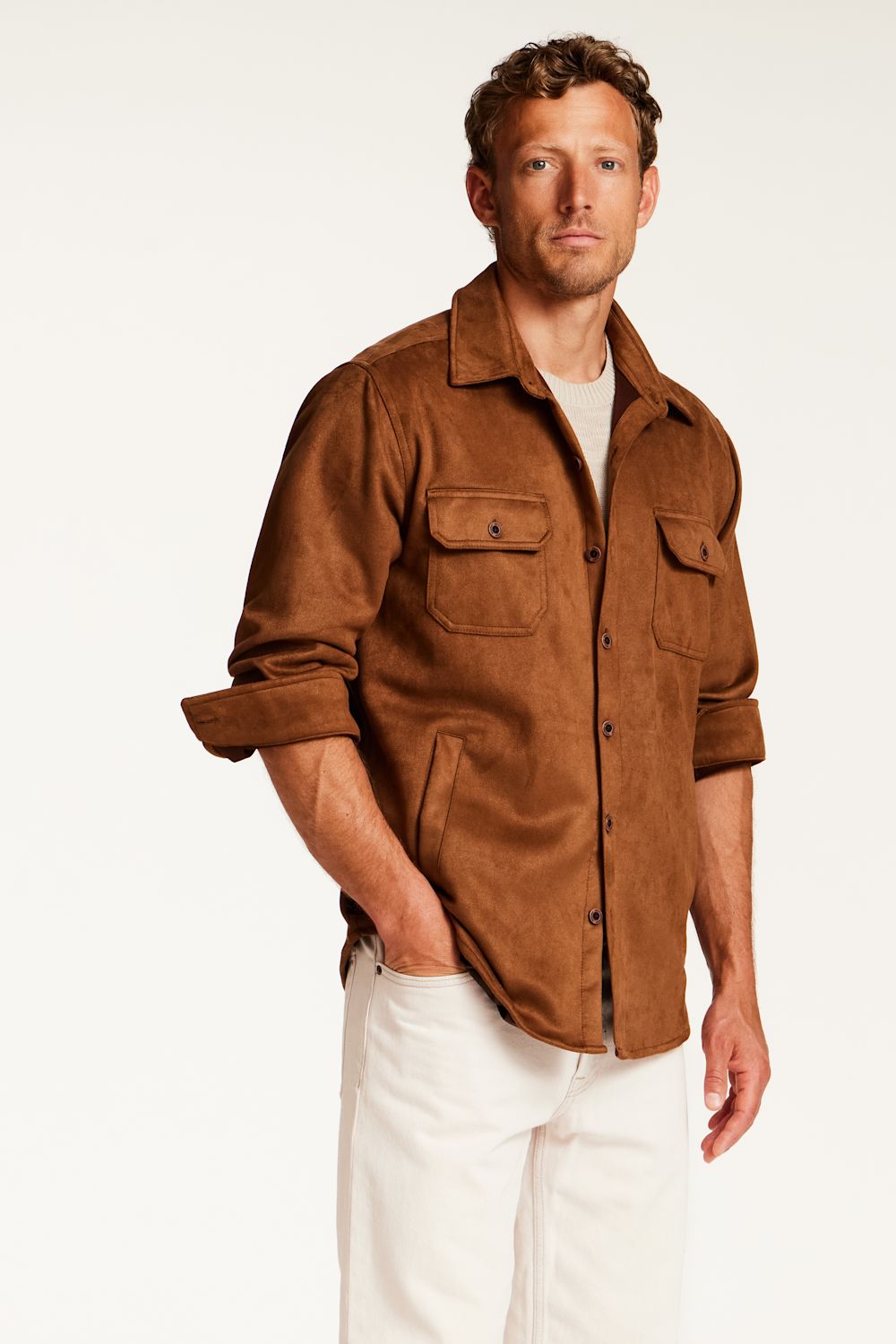A man wearing a brown suede-like button-up shirt with chest flap pockets, worn open over a white t-shirt and white pants, photographed against a clean white studio background.