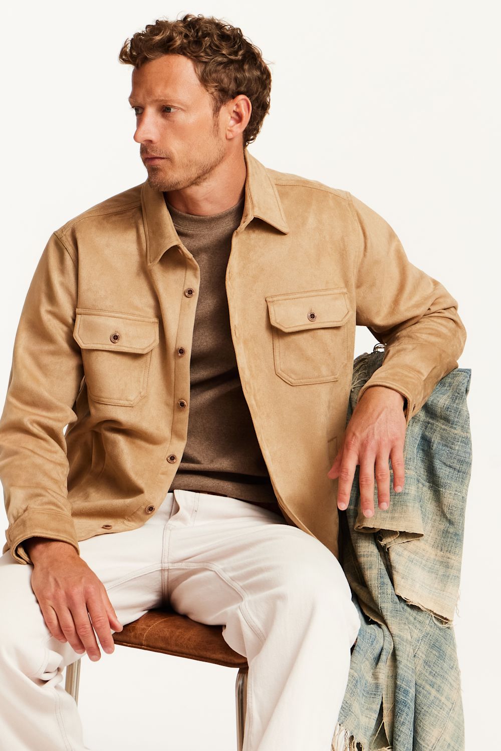 A man wearing a tan suede-like button-up shirt jacket over a brown t-shirt and white trousers, seated on a wooden chair with a blue-gray patterned flannel draped over the back of the chair, against a plain white studio background.