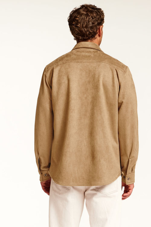 A man viewed from the back wearing a tan faux suede long-sleeve button-up shirt and off-white trousers, set against a plain white studio background.