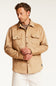 A man wearing a tan suede-like button-up shirt jacket with two chest flap pockets, styled over a brown t-shirt and white pants, photographed against a clean white studio background.