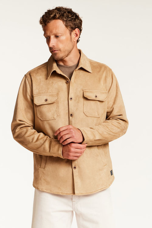 A man wearing a tan suede-like button-up shirt jacket with two chest flap pockets, styled over a brown t-shirt and white pants, photographed against a clean white studio background.