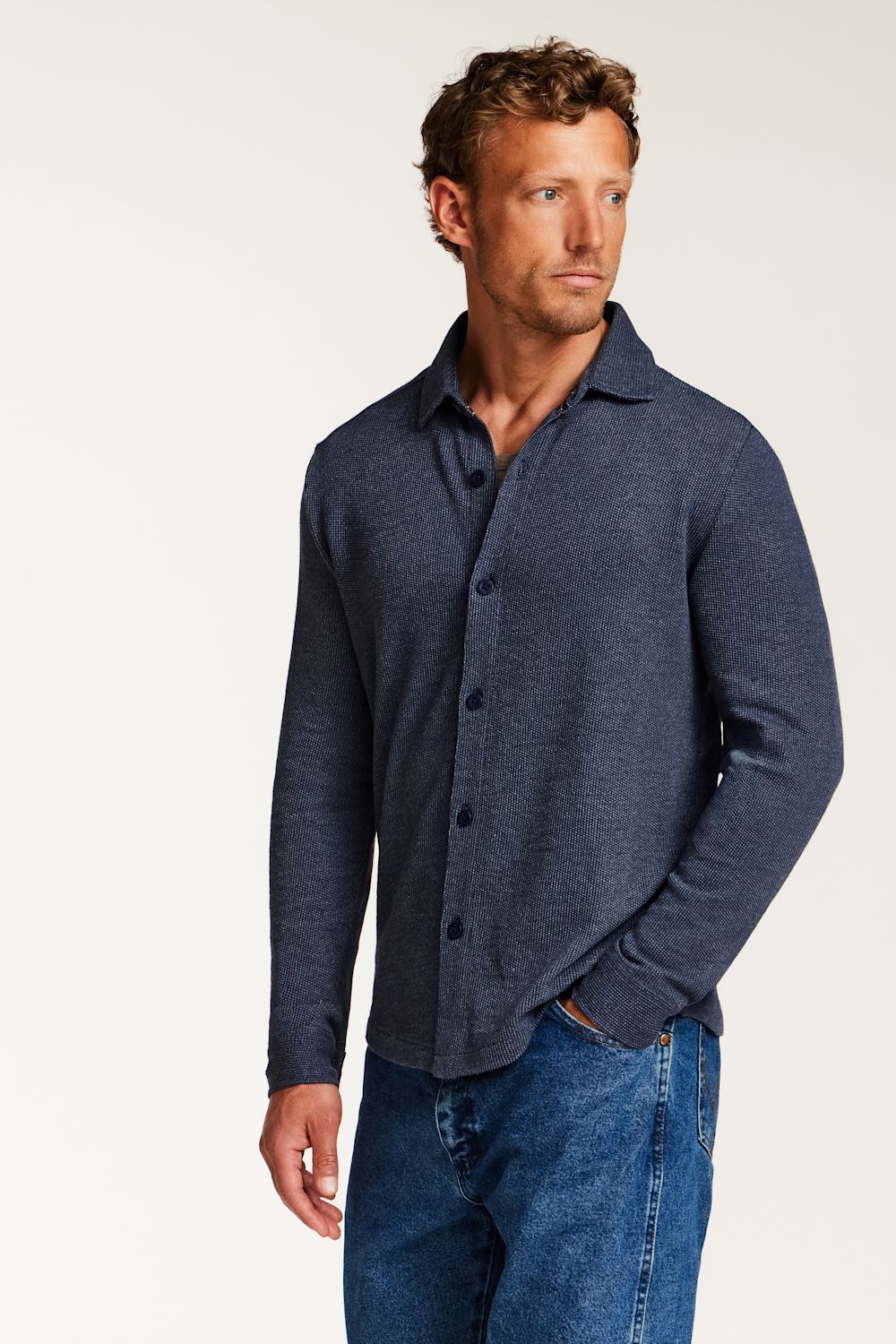 A man wearing a dark blue heathered long-sleeve button-up shirt and blue jeans, standing against a plain off-white studio background.