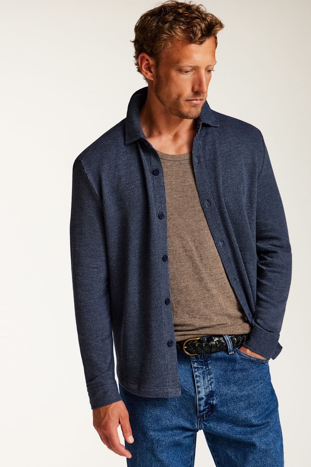 A man wearing a navy blue button-up knit overshirt over a taupe t-shirt and blue jeans, photographed against a clean off-white studio background.