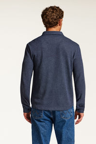 A man viewed from the back wearing a dark heathered blue long-sleeve polo shirt and blue jeans, set against a plain light background.