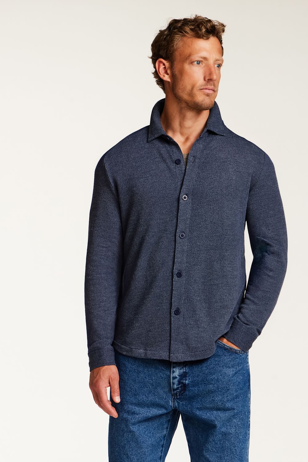 A man wearing a dark navy blue long-sleeve knit button-up shirt and blue jeans, standing against a plain light background.