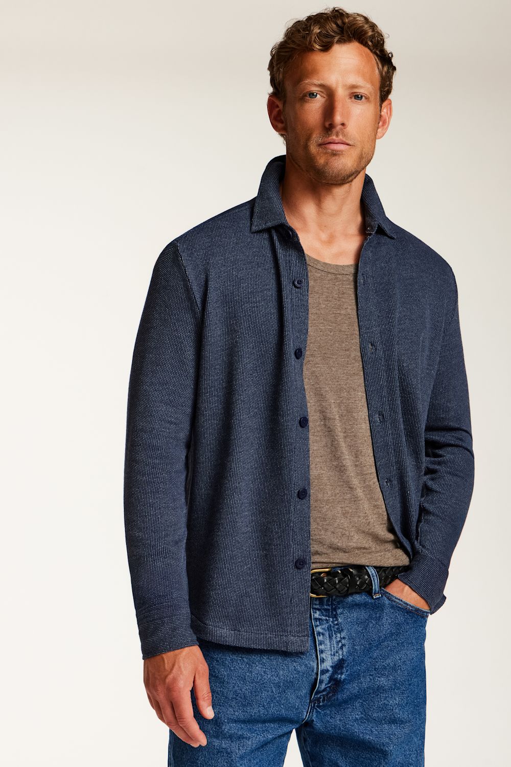 A man wearing a dark blue button-up shirt over a taupe t-shirt and blue jeans, standing against a plain light background.