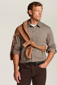 A man wearing a brown and beige checkered button-down shirt, dark brown trousers, and a brown knit sweater tied around his shoulders, photographed against a light gray studio background.