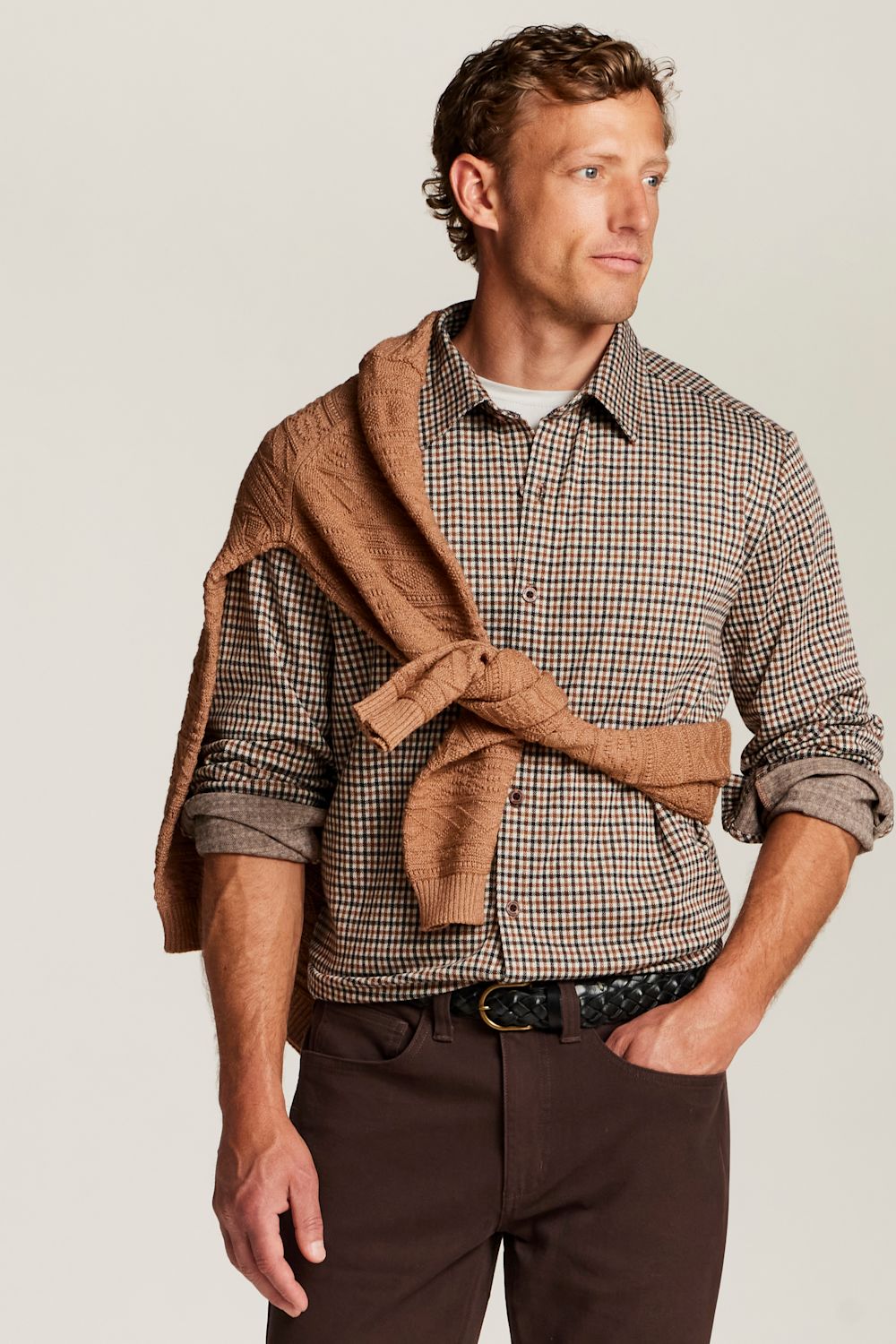 A man wearing a brown and beige checkered button-down shirt, dark brown trousers, and a brown knit sweater tied around his shoulders, photographed against a light gray studio background.