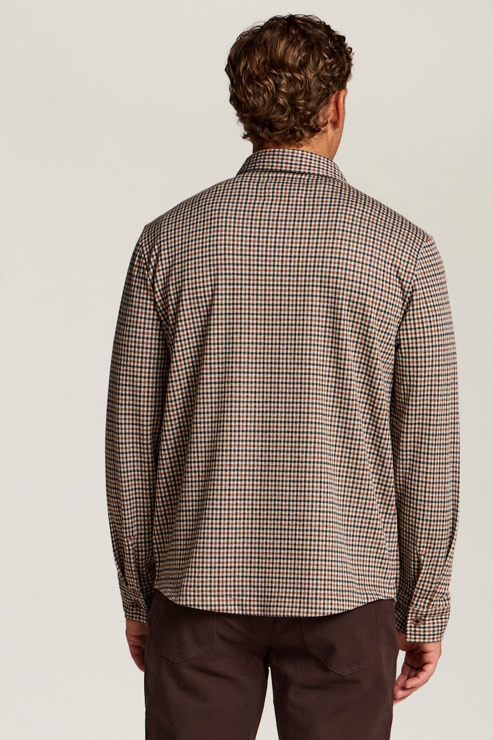 A man viewed from the back wearing a brown and beige checkered long-sleeve shirt and dark brown trousers, set against a plain light gray studio background.