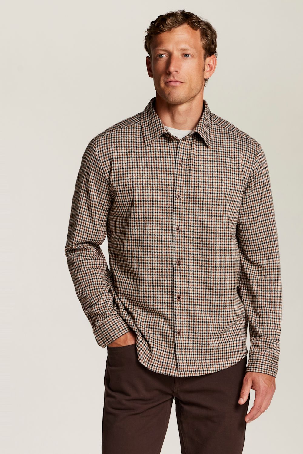 A man wearing a brown and beige checkered long-sleeve button-down shirt with dark brown trousers, standing against a light gray studio background.