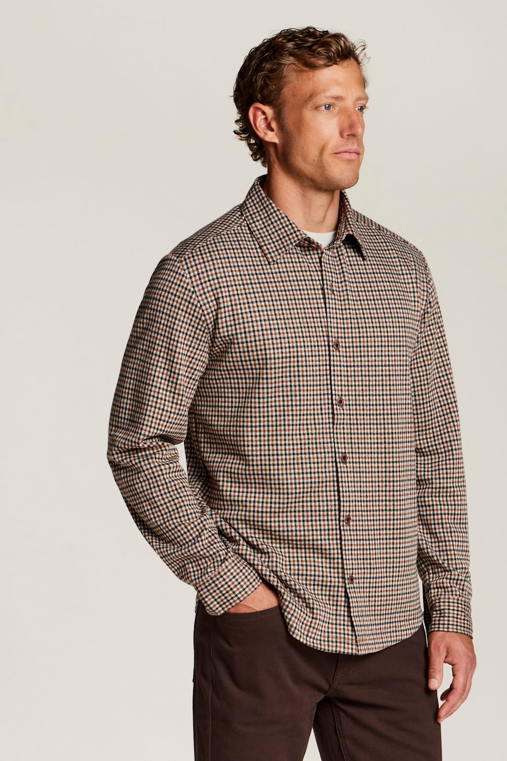 A man wearing a brown and beige gingham long-sleeve button-down shirt, paired with dark brown trousers, standing against a light gray studio background.