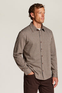 A man wearing a brown and beige gingham long-sleeve button-down shirt, paired with dark brown trousers, standing against a light gray studio background.