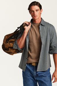 A man wearing a black and white gingham overshirt over a brown crewneck sweater, blue jeans, and holding a brown duffel bag, photographed against a light gray studio background.