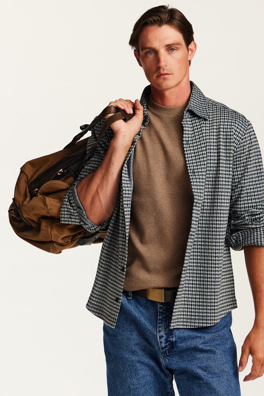 A man wearing a black and white gingham overshirt over a brown crewneck sweater, blue jeans, and holding a brown duffel bag, photographed against a light gray studio background.