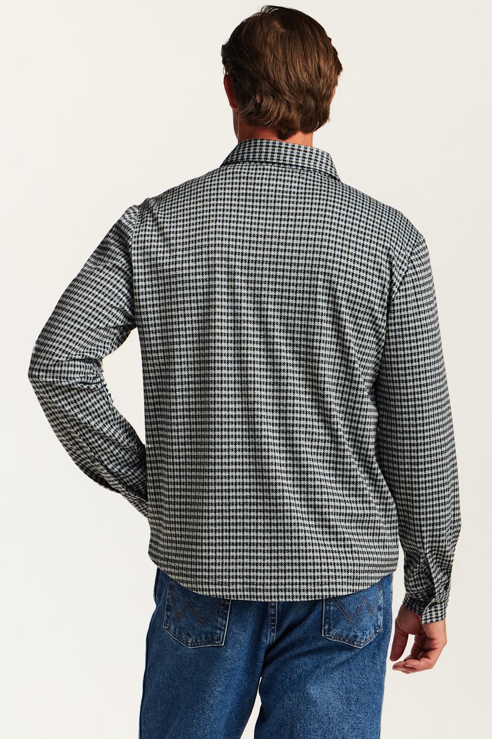 A man viewed from behind wearing a long-sleeve button-up shirt with a black and white micro-check pattern, paired with blue jeans, against a plain off-white background.