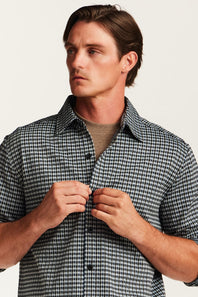 A man wearing a short-sleeve black and white gingham check shirt over a beige crewneck t-shirt, buttoning the collar and front of the shirt against a plain off-white background.