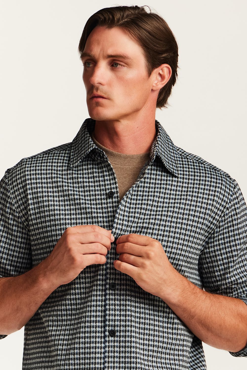 A man wearing a short-sleeve black and white gingham check shirt over a beige crewneck t-shirt, buttoning the collar and front of the shirt against a plain off-white background.