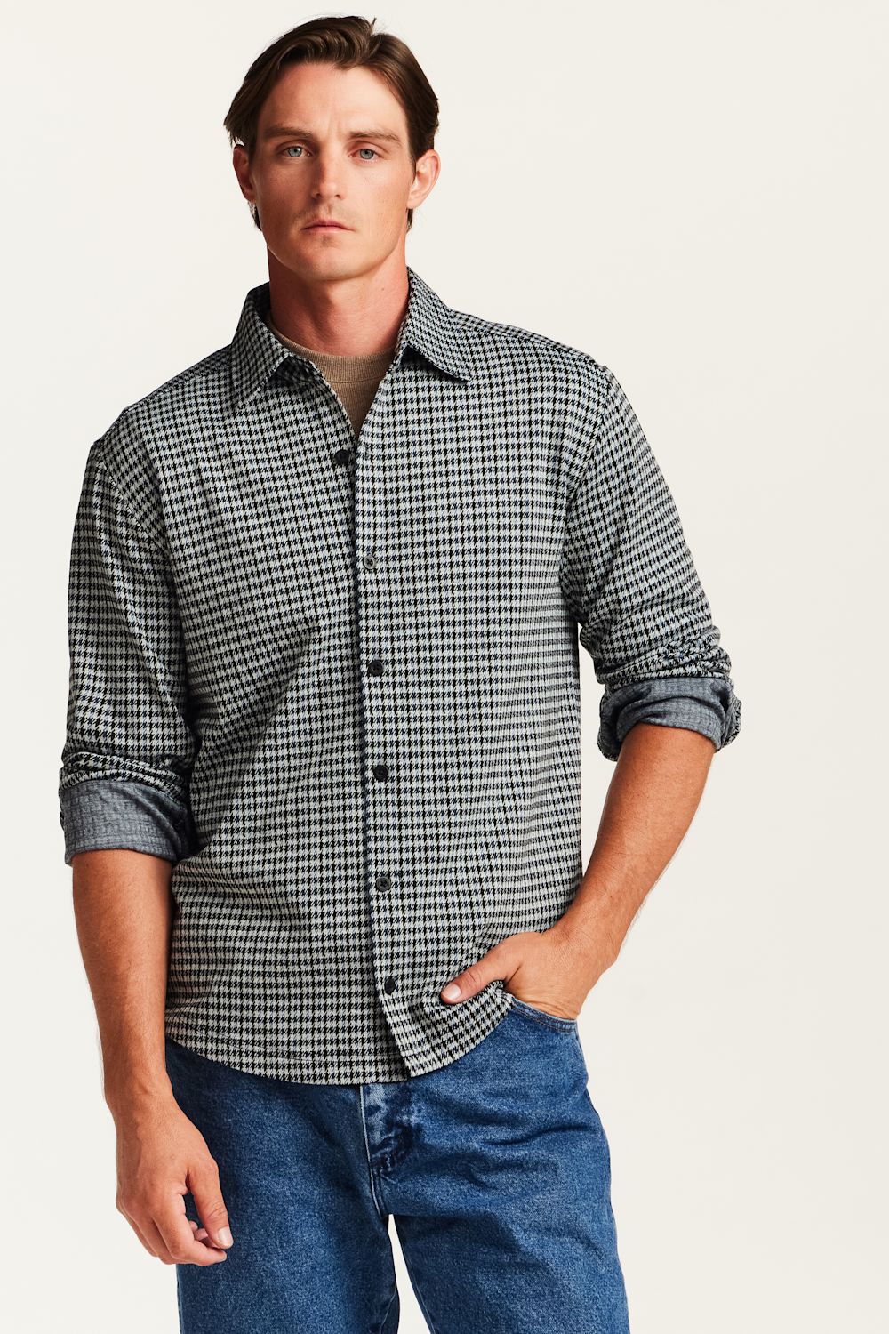 A man wearing a long-sleeve button-down shirt with a small black and white houndstooth pattern, paired with blue jeans, against a light off-white background.
