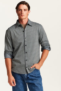A man wearing a long-sleeve button-down shirt with a small black and white houndstooth pattern, paired with blue jeans, against a light off-white background.