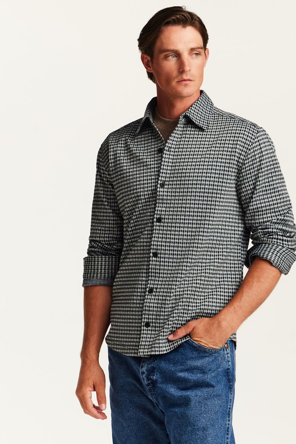 A man wearing a black and white micro-check patterned button-down shirt with rolled-up sleeves, paired with blue jeans, against a plain off-white background.