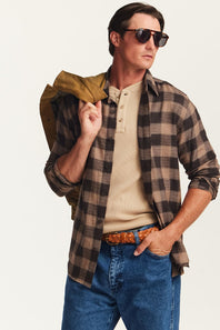 A man wearing a brown and black plaid button-down shirt over a beige henley tee, blue jeans, and sunglasses, holding a brown jacket over his shoulder against a plain light background.