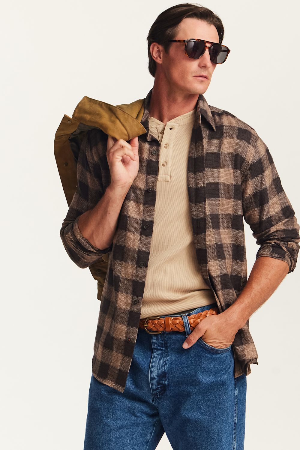 A man wearing a brown and black plaid button-down shirt over a beige henley tee, blue jeans, and sunglasses, holding a brown jacket over his shoulder against a plain light background.
