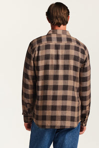 A man viewed from the back wearing a brown and beige plaid flannel shirt and blue jeans, set against a plain off-white background.