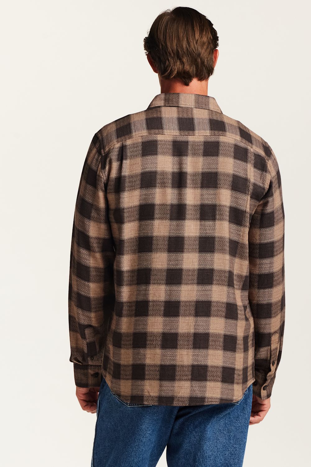 A man viewed from the back wearing a brown and beige plaid flannel shirt and blue jeans, set against a plain off-white background.