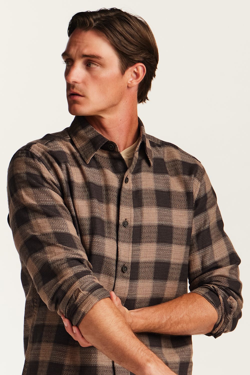 A man wearing a brown and beige plaid button-down flannel shirt, shown from the chest up against a plain off-white background.